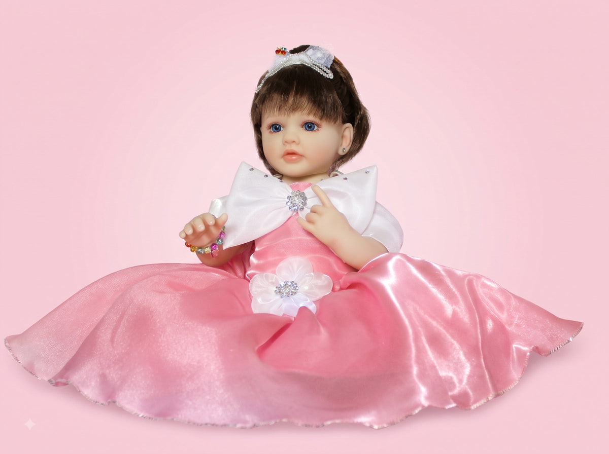 A realistic baby doll with brown hair and blue eyes, wearing a shiny pink satin dress with a large white bow at the collar and a silver tiara, posed against a solid pink background.