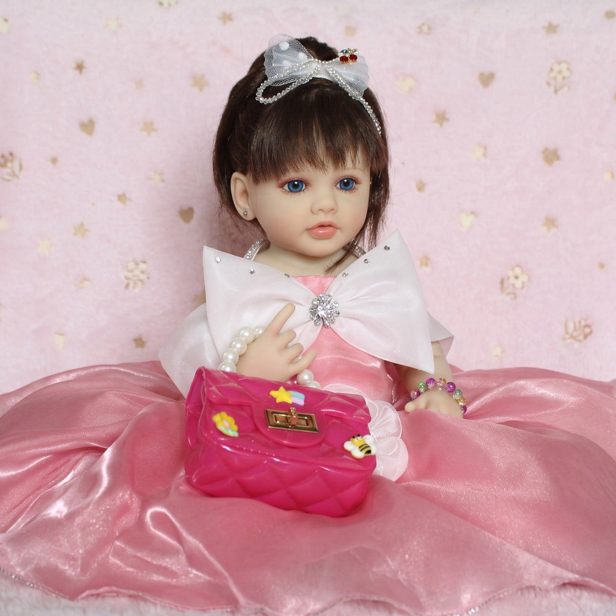 A doll with brown hair and blue eyes wearing a pink dress with a large white bow, seated on a pink patterned blanket and holding a pink quilted handbag.